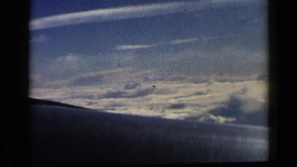 1960 8mm view from an airplane window with the wing along the lower frame and sunlit cloud layers beyond.