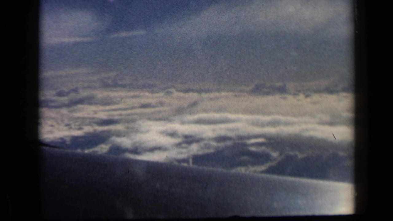 Airplane wing seen through a dark window frame above a dense layer of clouds.