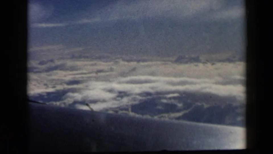 Aerial 8mm view from a plane window, showing the wing edge against layered clouds during a 1960 travel shot.