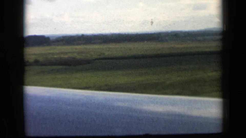 1960 8mm airplane-window view of green rural fields below, with the wing foreground and soft horizon in the distance.