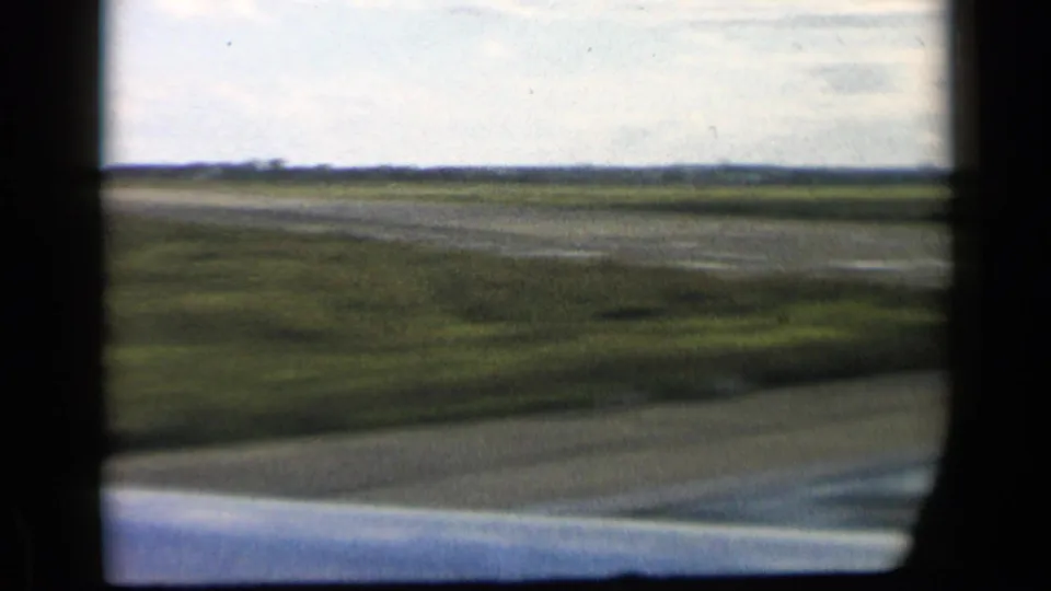 1960 8mm vehicle-perspective view over open fields, with a low horizon, pale sky, and dark window-like framing.