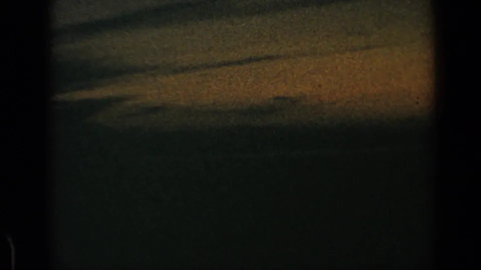 1960 8mm footage darkens as the moving vehicle passes low fields or roadside ground, with heavy shadow at the bottom.