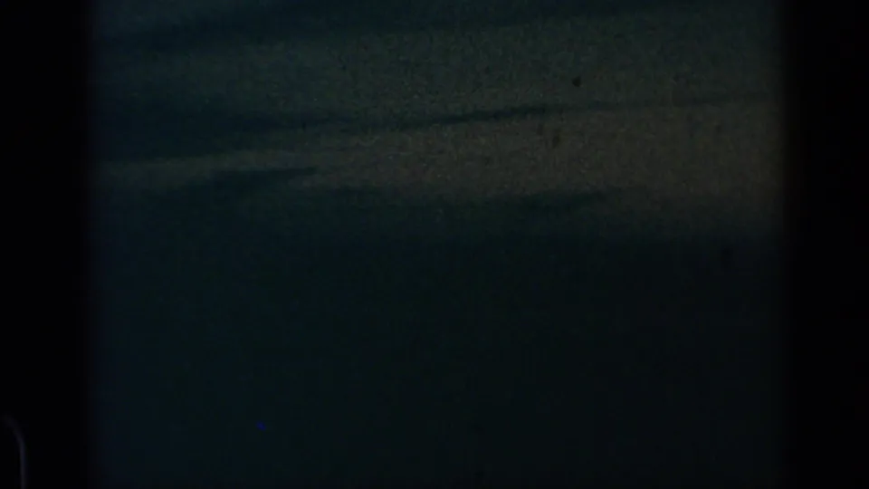 Dark 1960 8mm frame of evening clouds from an aircraft, marked by grain, low exposure, and window-edge shadows.