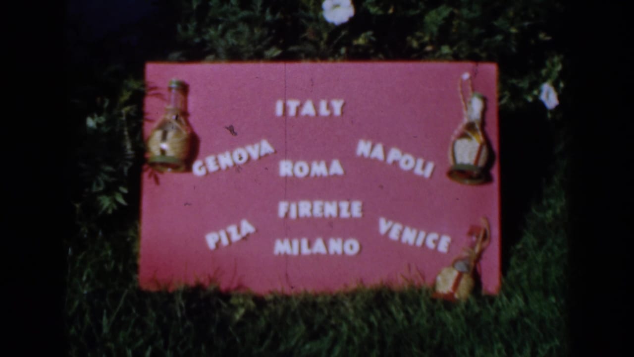 Pink outdoor sign with white Italian city names and bottle ornaments on grass.