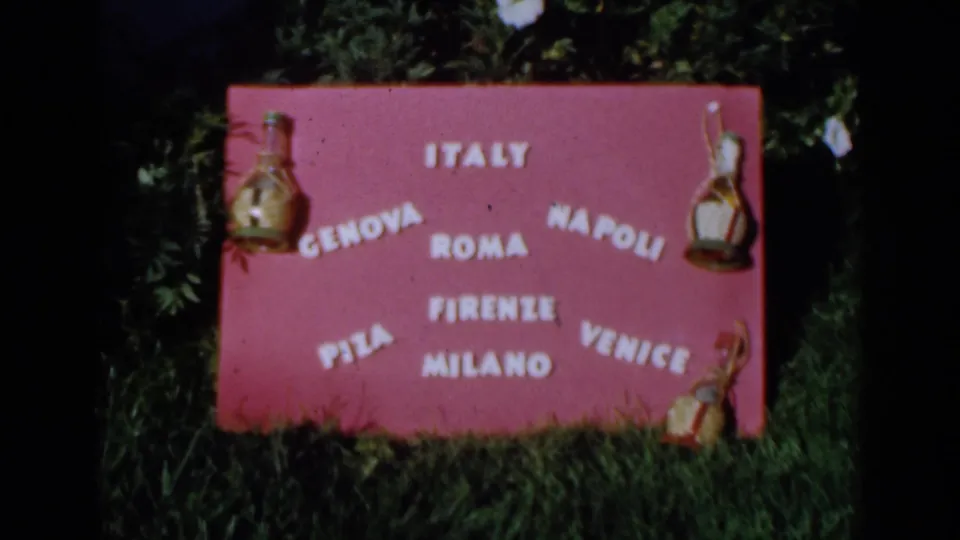 1960 8mm frame showing the same pink Italian cities sign outdoors, bordered by grass and leafy foliage.
