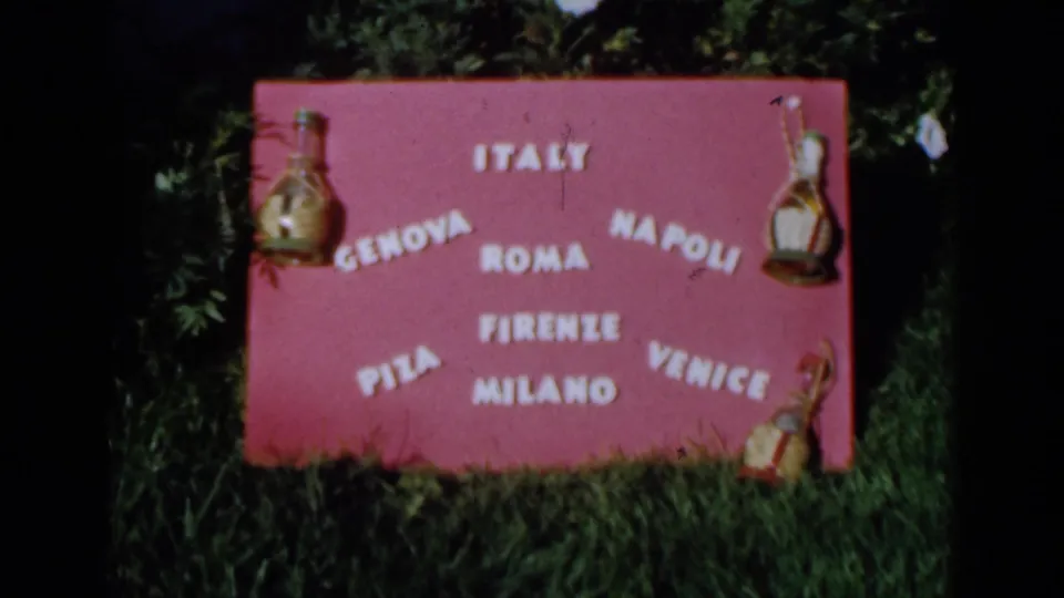 1960 8mm frame with a handheld view of the pink sign, white city lettering, and bottle ornaments in an outdoor setting.