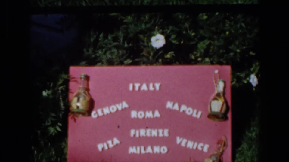 Final 1960 8mm frame shows the Italy-themed city sign slightly soft, with green foliage above and grass below.