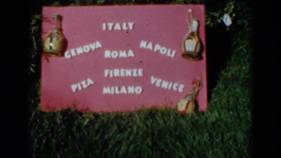 1960 Venice home-movie still of an Italy-themed pink sign naming several cities beside grass and greenery.