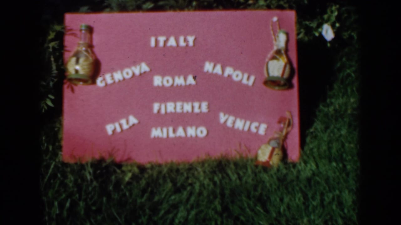 Pink handmade sign on grass with white Italian city names and bottle ornaments.