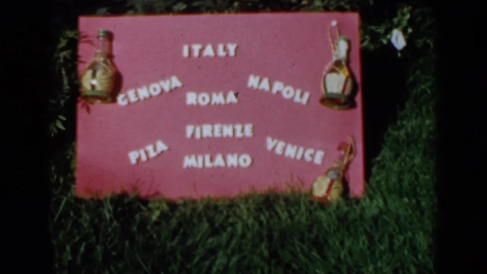 1960 Venice, Italy home-movie frame showing a pink handmade sign listing Italian cities, decorated with small bottle-shaped ornaments on grass.