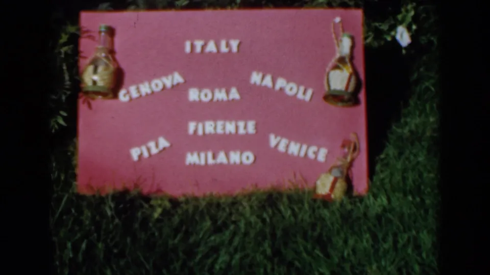 1960 Venice, Italy frame of a handmade pink sign reading Italy and listing cities including Roma, Napoli, Firenze, Milano, and Venice.