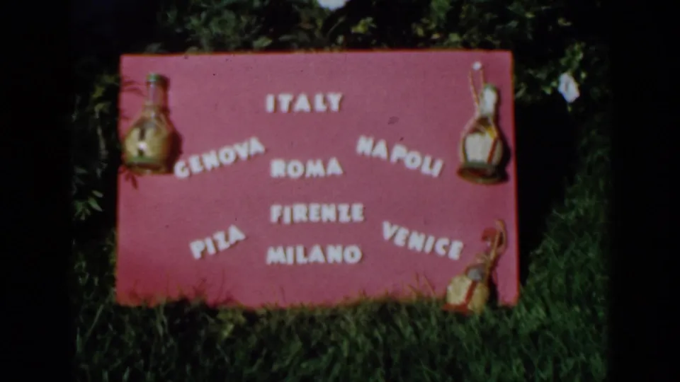 1960 Venice, Italy home-movie frame showing the pink travel sign from a slightly shifted angle, with white city names and small ornaments.