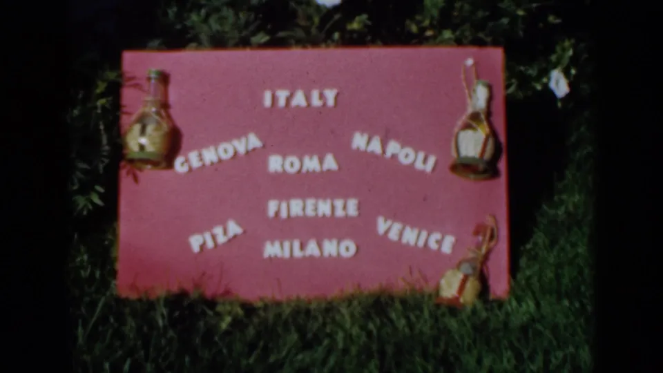 1960 Venice, Italy frame showing the handmade pink sign with Italian city names, bottle ornaments, green grass, foliage, and visible film borders.