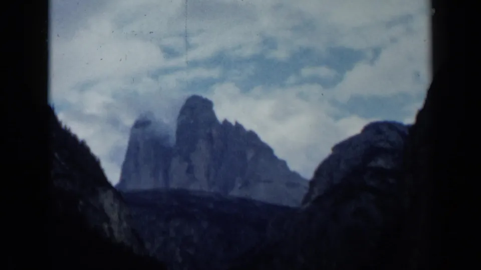 1960 8mm Italian travel footage showing distant rocky peaks under clouds, seen through dark silhouetted foreground rock.