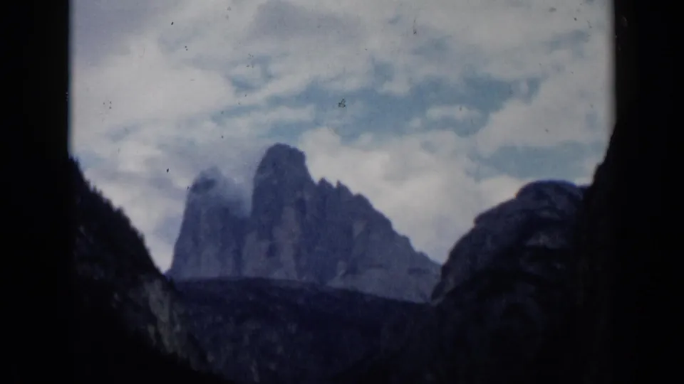 1960 8mm travel shot of distant rocky peaks and cloud cover, framed by dark foreground cliffs in an Italian mountain landscape.