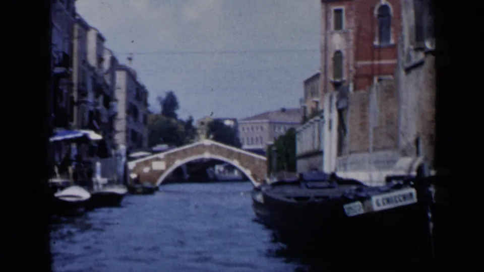 Venice, Italy canal view from 1960 home-movie footage, with an arched bridge, moored boats, and buildings along the water.