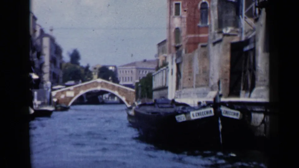 1960 Venice canal footage shows an arched pedestrian bridge, narrow waterway, moored boats, and weathered buildings.
