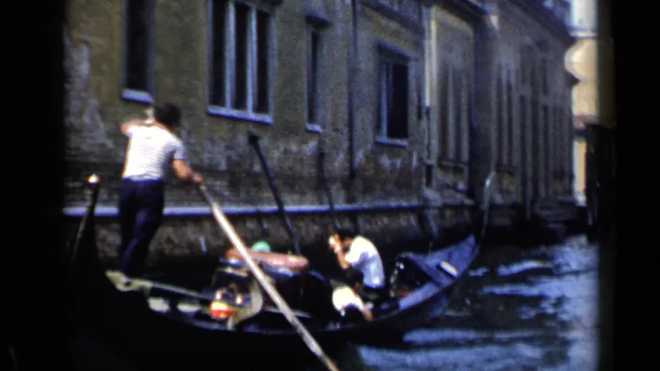 A gondolier in a white shirt poles a small boat with passengers along a Venice canal in 1960 home-movie footage.