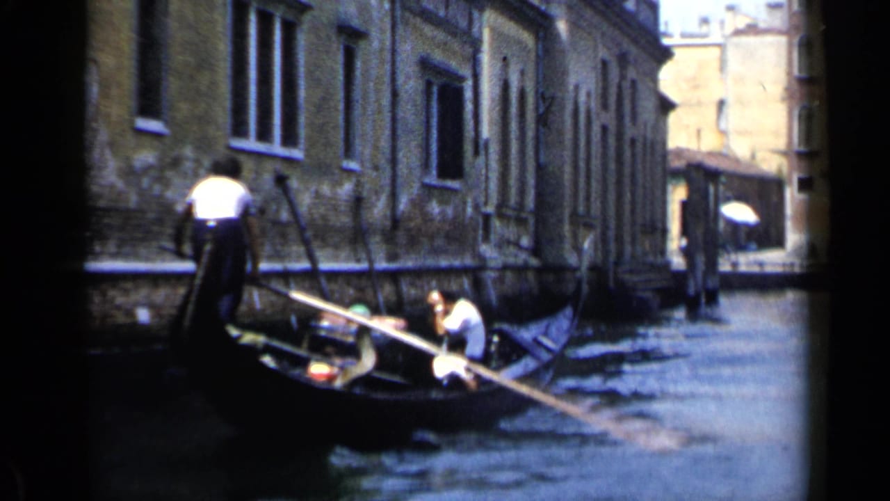 1960 Venice home-movie frame showing a gondolier guiding a gondola through a narrow canal between masonry buildings.