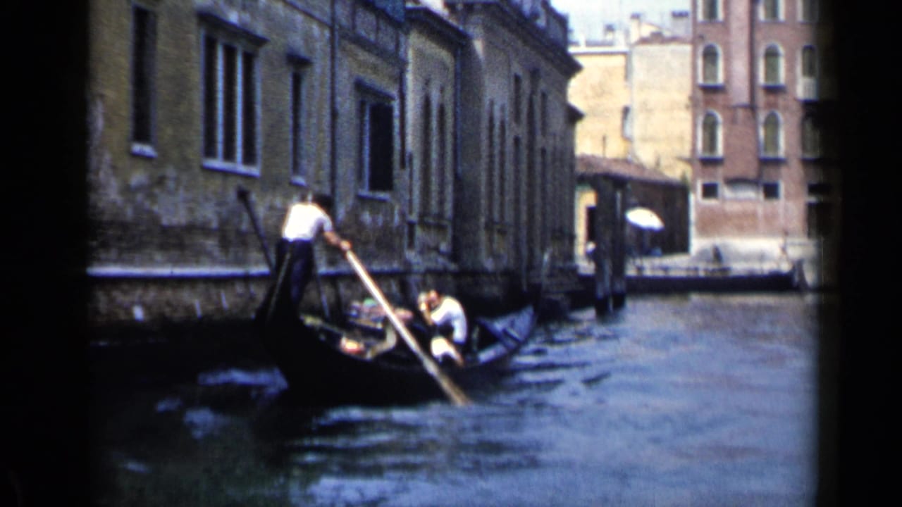 A gondolier in a white shirt steers a black gondola through a Venice canal, with passengers partly visible in the boat.