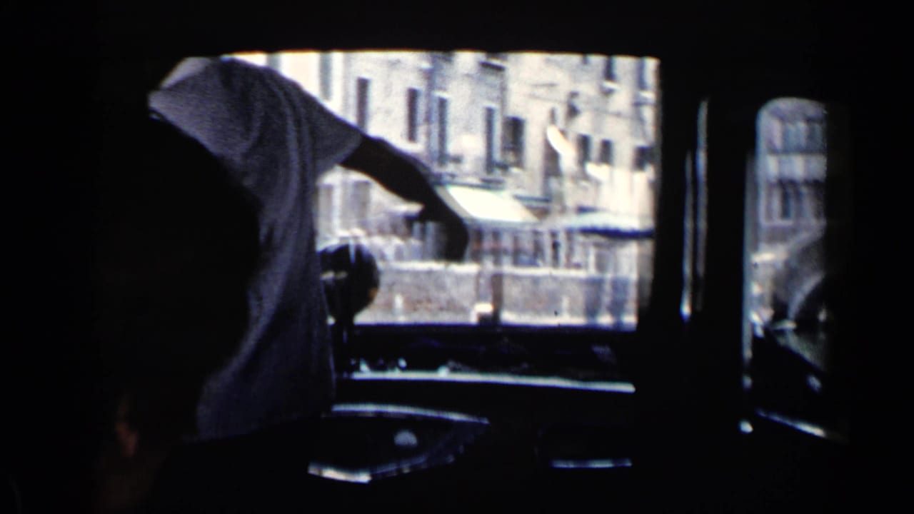 Dark boat interior with a man leaning near the window as canal buildings pass outside.