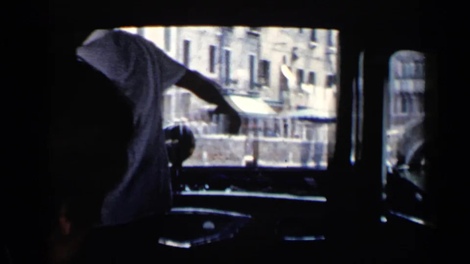 Inside a Venice canal boat in 1960, a passenger in a white shirt leans near the window while buildings and water are visible outside.