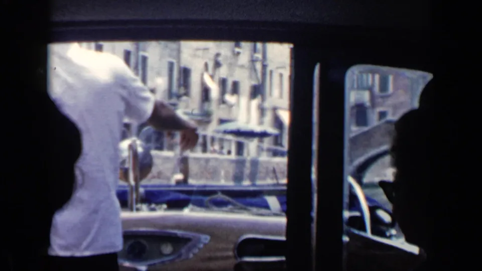 A tourist aboard a Venice canal boat points through the window toward waterside buildings and moored boats in the supplied 1960 footage.