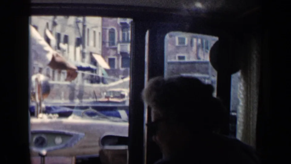 From a shadowed boat interior, a pointing arm extends toward the Venice canal while a seated passenger is silhouetted below the window.