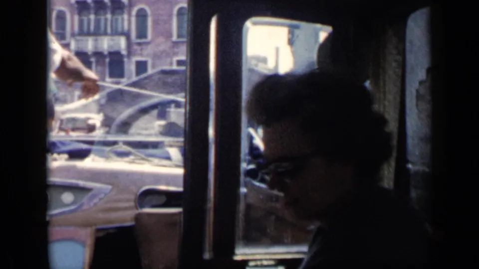 A smiling woman in sunglasses sits in the boat interior as Venice canal architecture and another boat appear through the window.
