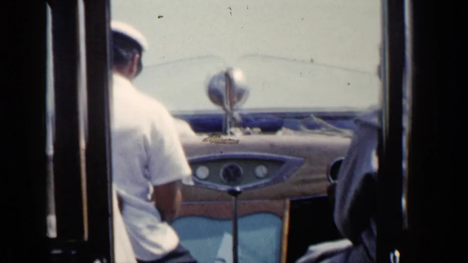 1960 Venice boat operator in a white shirt and cap seated near the helm, with the windshield and dashboard in view.