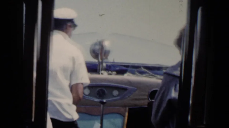 1960 Venice canal boat view with a white-capped operator at the helm and another passenger partly visible in the foreground.