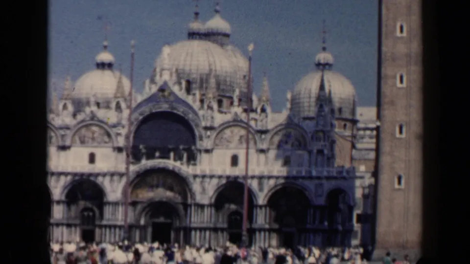 1960 Venice view of St. Mark's Basilica with ornate arches, domes, a bell tower edge, and tourists gathered in front.
