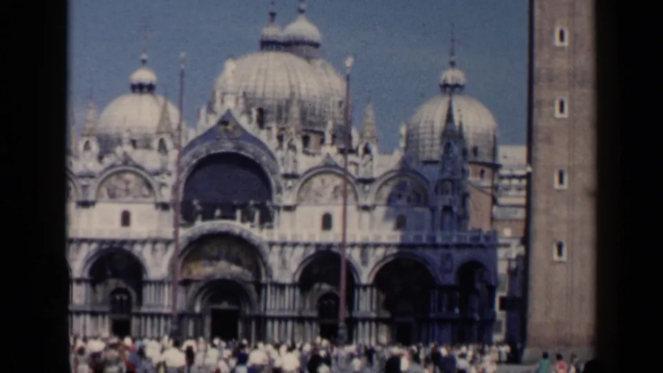 Venice 1960 home-movie frame of St. Mark's Basilica, showing domes, ornate arches, tower edge, and a crowd in the square.