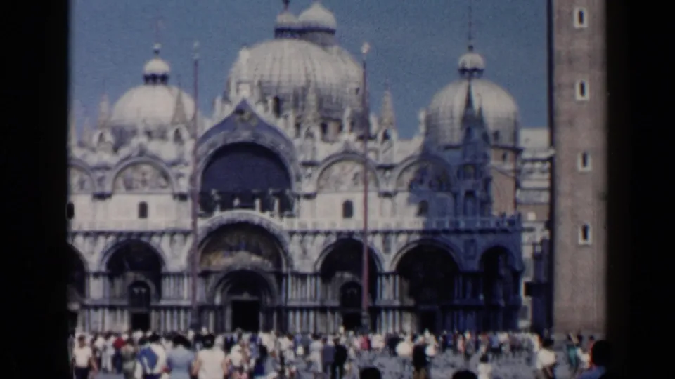 1960 8mm view of St. Mark's Basilica in Venice, with its domed facade, arched portico, campanile edge, and visitors below.