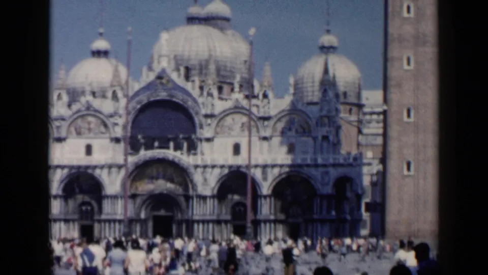 Wide 1960 Venice frame showing St. Mark's Basilica, the nearby campanile edge, arched facade details, and a tourist crowd at ground level.