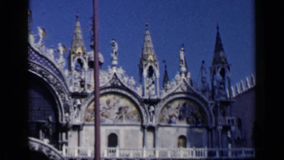 1960 Venice 8mm frame showing lower facade arches of St. Mark's Basilica, mosaic panels, spires, statues, and partial shadow.