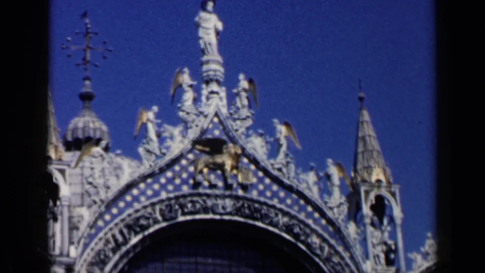 1960 8mm Venice view of St Mark's Basilica facade, showing sculptural figures, patterned arch detail, and a winged lion motif.