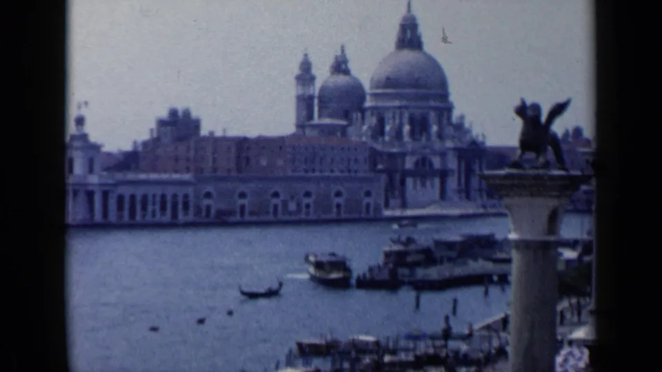 1960 8mm Venice view across the canal toward Santa Maria della Salute, with water traffic and a winged lion column at right.