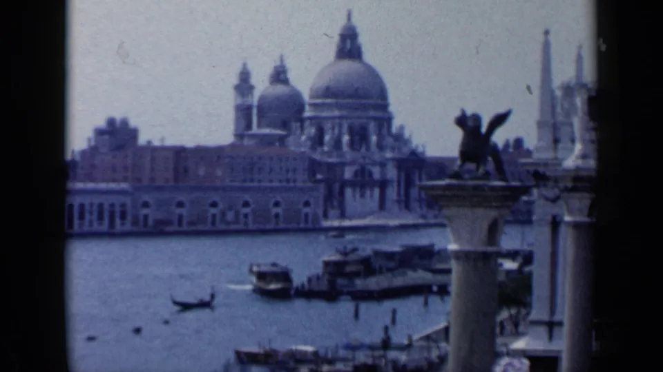 1960 8mm Venice frame showing Santa Maria della Salute across the canal, gondolas below, and a winged lion column near the right edge.