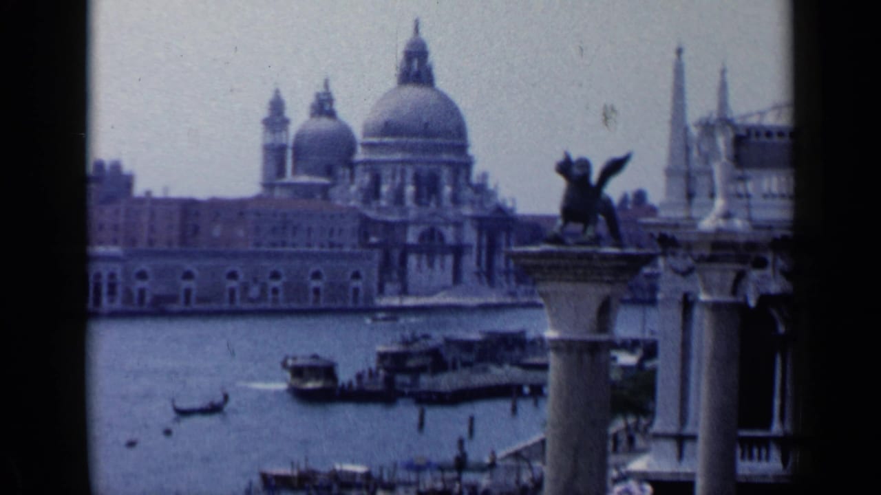 1960 Venice home-movie frame showing Santa Maria della Salute across the water, with a winged lion column, gondolas, and motorboats in view.