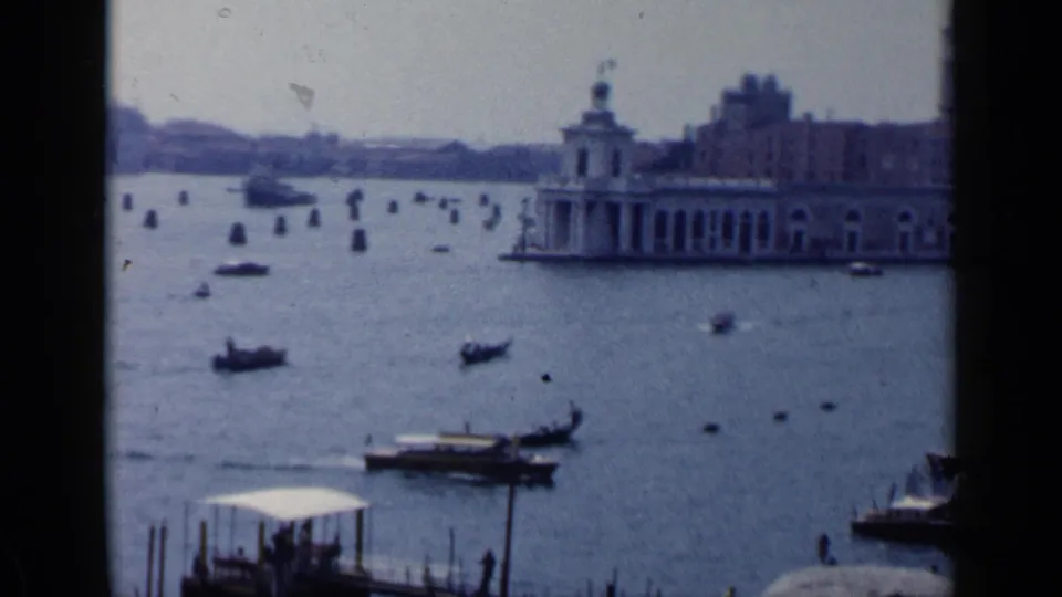 1960 Venice wide view showing open water, gondolas, motorboats, rows of mooring posts, and historic waterfront buildings in soft 8mm color.