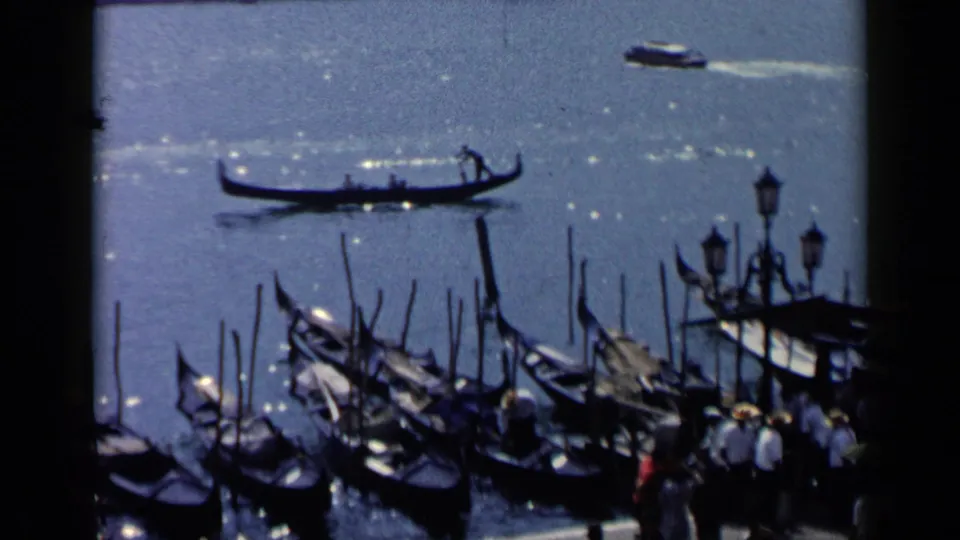 1960 Venice 8mm frame showing gondolas on shimmering water, moored boats, dockside figures, and lamps near the canal.