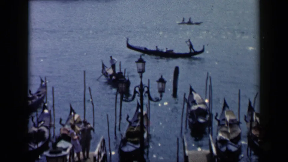 1960 Venice view of a gondola on blue canal water with clustered moored boats, mooring poles, lamps, and small figures below.