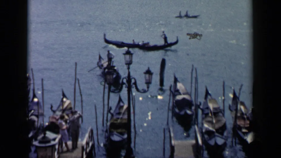 1960 Venice frame with moored gondolas, tall poles, waterfront lamps, pedestrians, and a gondola visible on the canal.