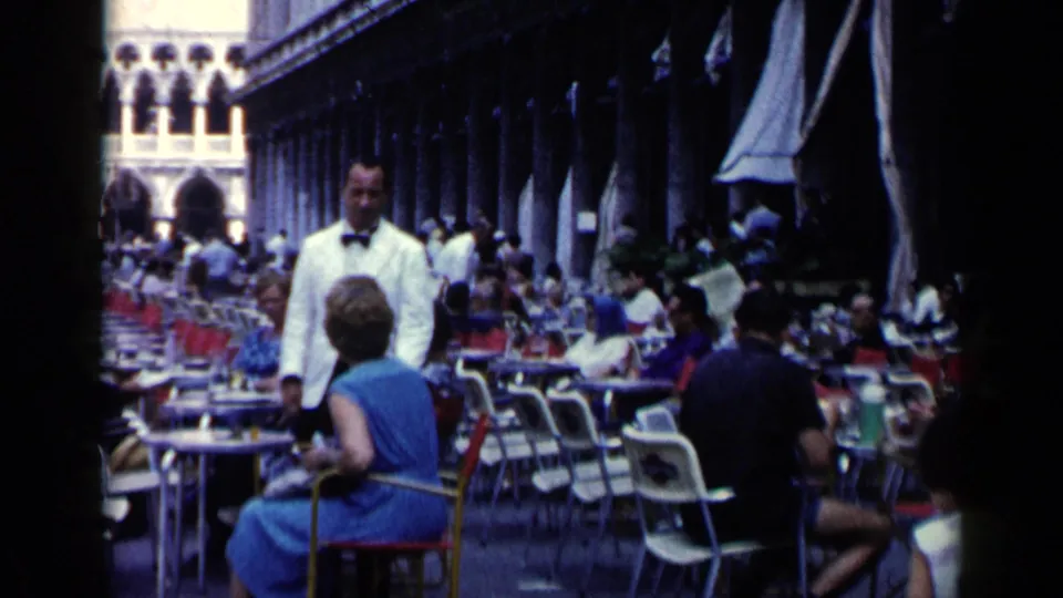 1960 Venice 8mm frame showing a busy outdoor café under arcades, with seated patrons, red chairs, and a waiter in a white jacket.