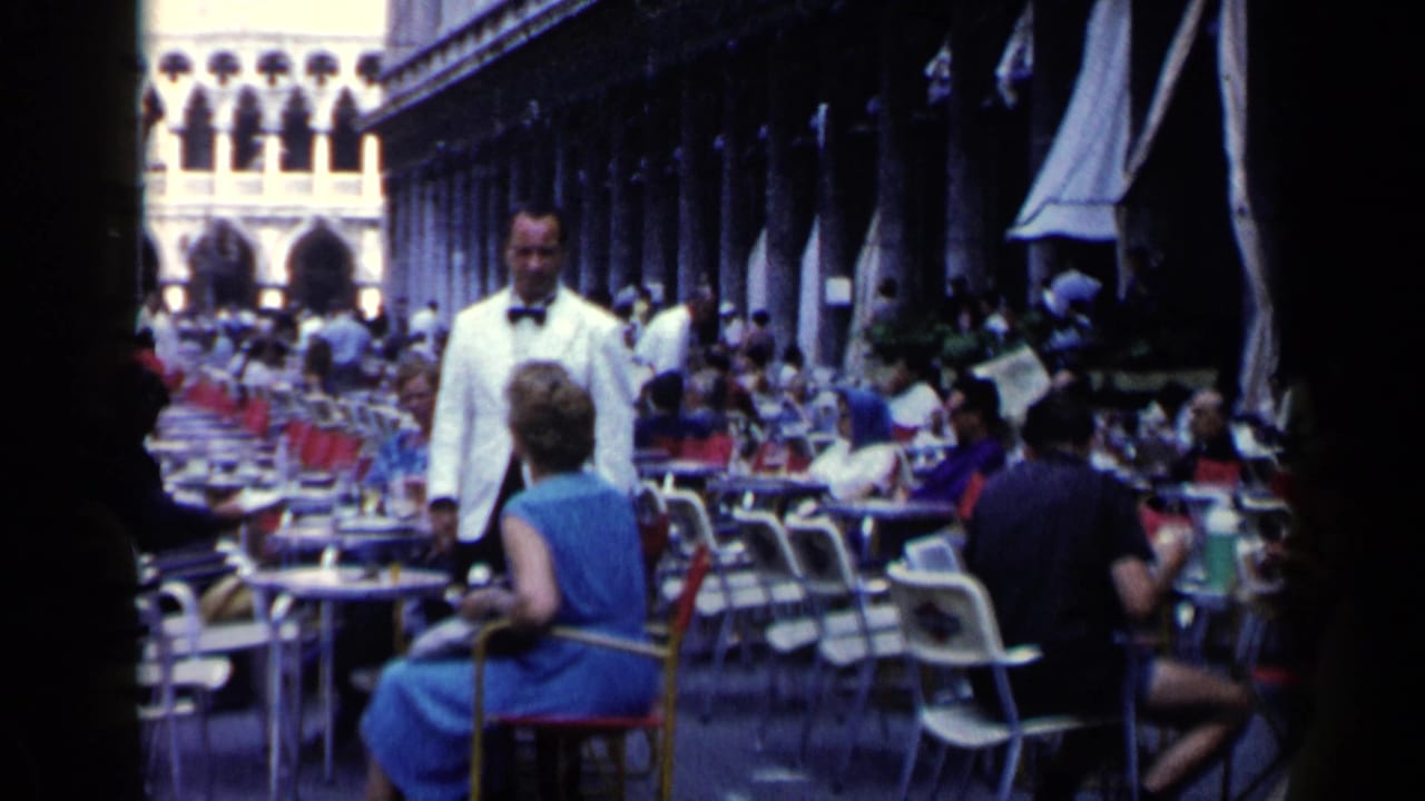 Waiter in a white jacket stands near a seated woman at a busy outdoor cafe.