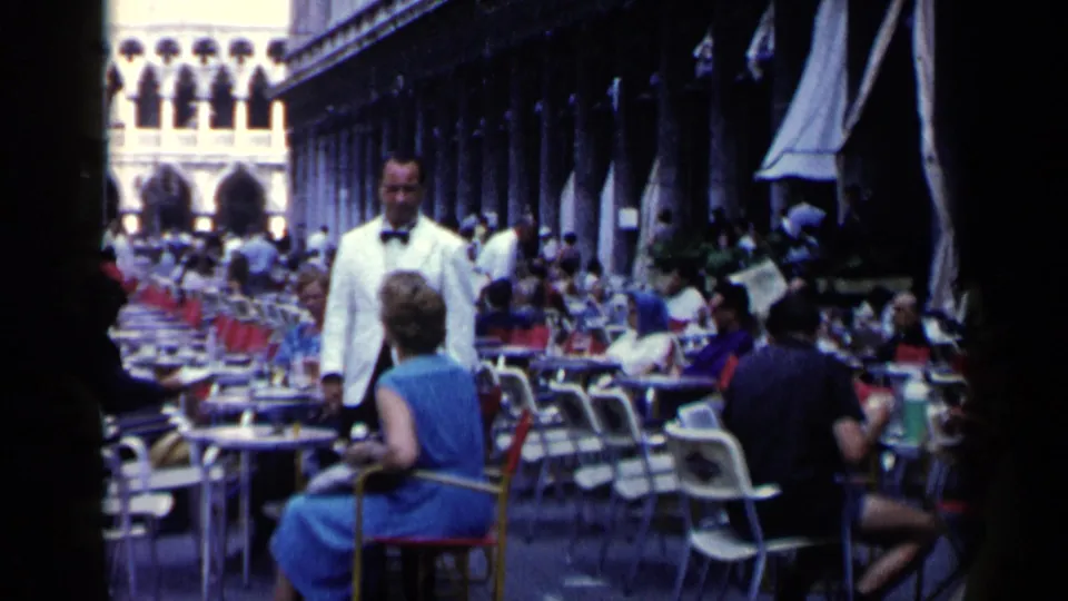1960 Venice outdoor cafe scene with a white-jacketed waiter, seated woman in blue, crowded tables, and shaded arcade.