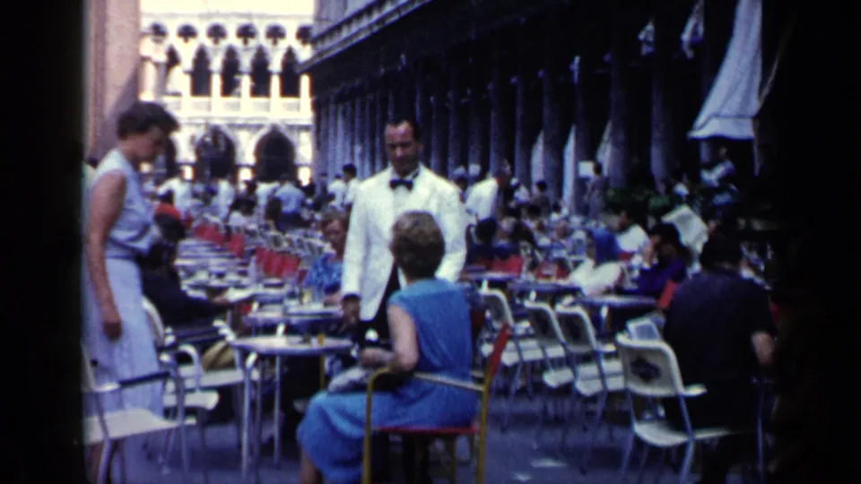 At a Venice cafe in 1960, a woman in a pale dress passes between tables as a waiter stands near seated patrons.