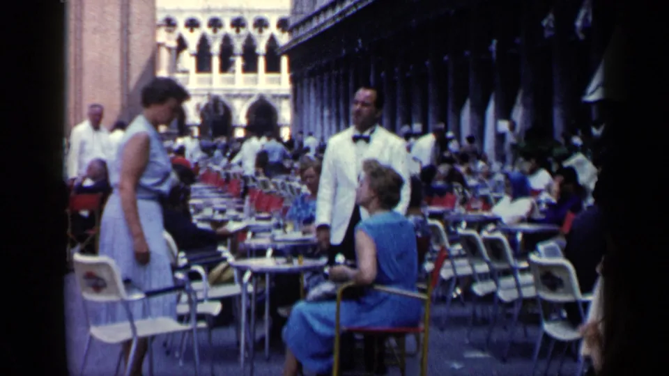1960 Venice terrace cafe with a white-jacketed waiter, standing woman, seated patron in blue, and rows of tables under an arcade.