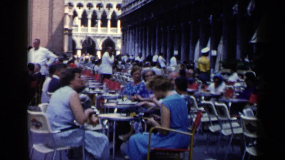 Wide 1960 Venice cafe view showing patrons seated at small tables under a dark arcade with bright arched architecture beyond.